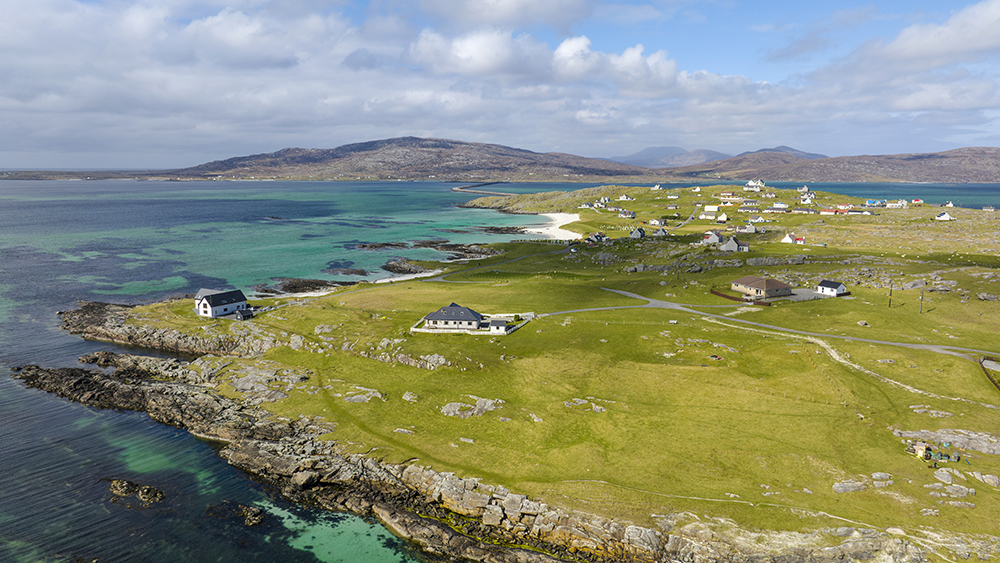 View from the sky of the isle of Eriskay, with turquoise water and white sand beach, and a few houses scattered on the land.